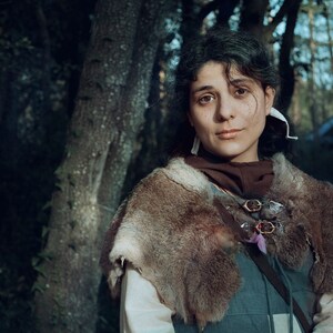 May include: A woman with dark hair and brown eyes is wearing a brown fur-trimmed cloak with a leather strap and metal buckles. She is standing in a forest setting.