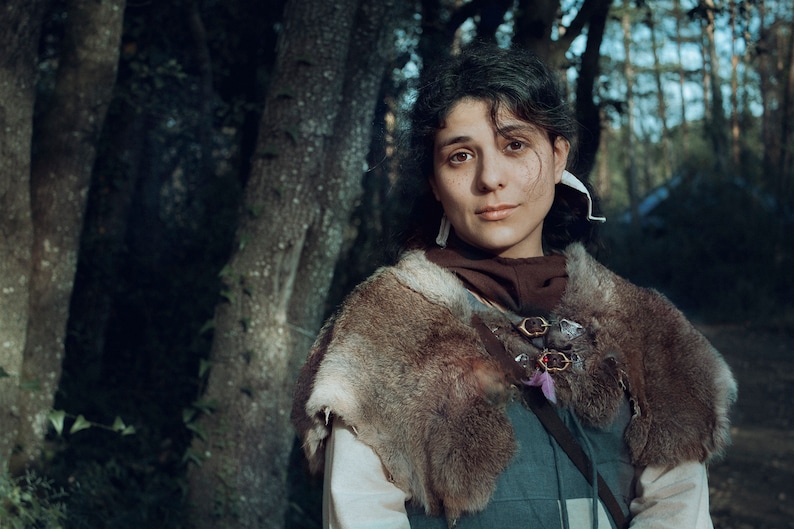May include: A woman with dark hair and brown eyes is wearing a brown fur-trimmed cloak with a leather strap and metal buckles. She is standing in a forest setting.