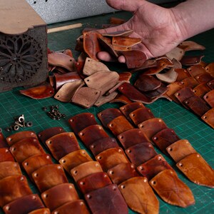 May include: A close-up of a collection of leather pieces, some are cut into shapes and others are still whole. The pieces are various shades of brown and are arranged on a green cutting mat. There are also small metal pieces scattered around the leather.