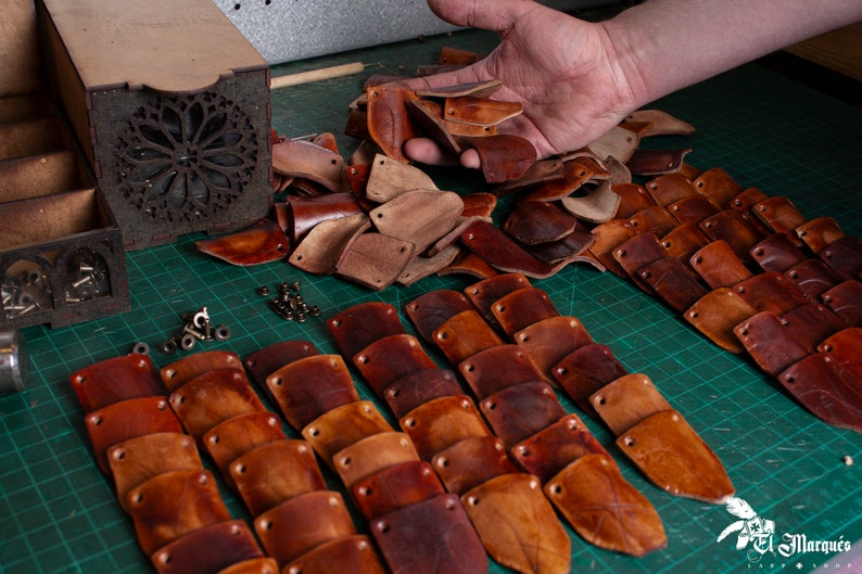 May include: A close-up of a collection of leather pieces, some are cut into shapes and others are still whole. The pieces are various shades of brown and are arranged on a green cutting mat. There are also small metal pieces scattered around the leather.
