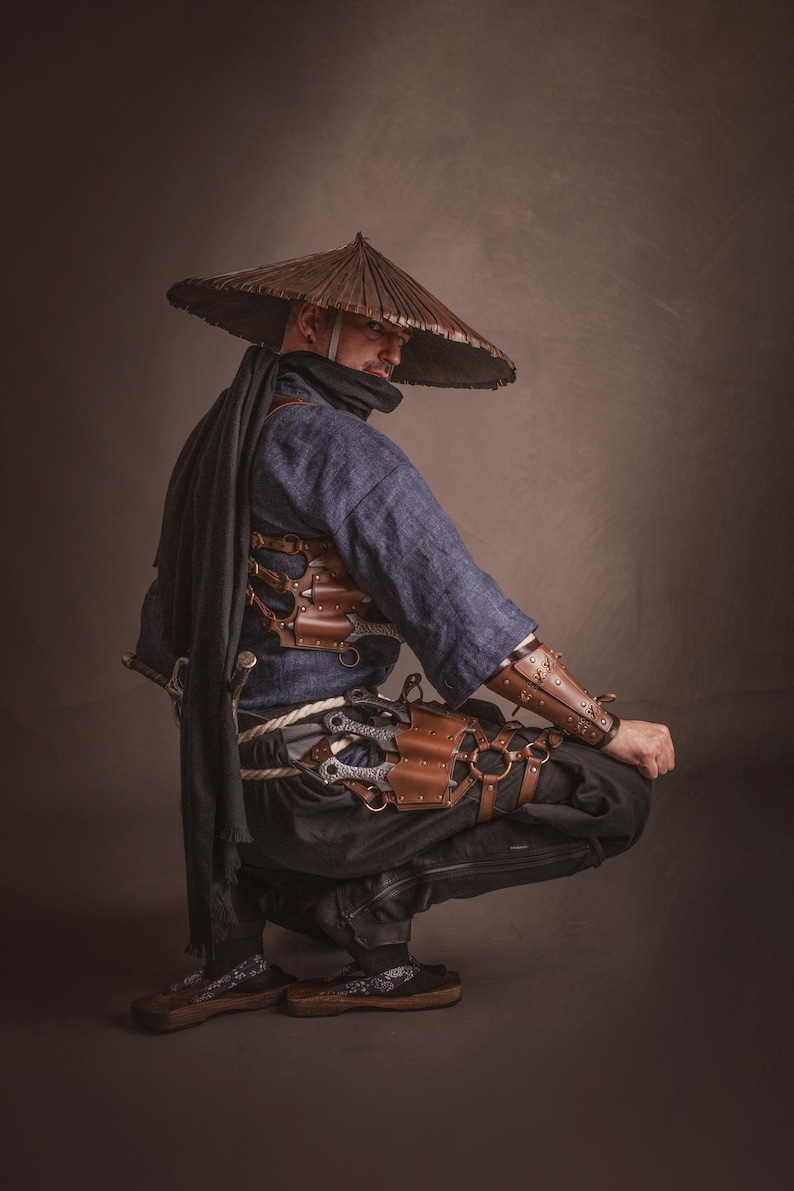 May include: A person wearing a blue shirt and brown leather armor with a wide-brimmed straw hat, holding a sword and crouching on a brown background.