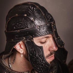K&ouml;nnte beinhalten: Schwarzer Lederhelm mit aufwendigen gepr&auml;gten Designs und Messingnieten. Der Helm hat ein Stirnband und Wangenplatten. Das Bild zeigt eine Nahaufnahme des Helms, die seine Handwerkskunst hervorhebt. Der Helm ist von einem LARP-Shop.