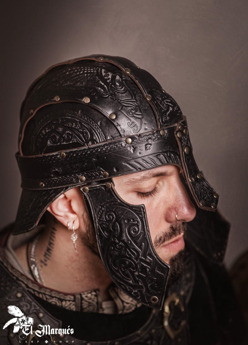 K&ouml;nnte beinhalten: Schwarzer Lederhelm mit aufwendigen gepr&auml;gten Designs und Messingnieten. Der Helm hat ein Stirnband und Wangenplatten. Das Bild zeigt eine Nahaufnahme des Helms, die seine Handwerkskunst hervorhebt. Der Helm ist von einem LARP-Shop.