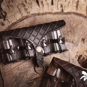 Peut inclure: &Eacute;tui en cuir noir avec un motif en losanges, contenant plusieurs petits flacons en verre. L'&eacute;tui comporte des &eacute;l&eacute;ments m&eacute;talliques d&eacute;coratifs et une fermeture avec un anneau. Le fond est une surface en bois.