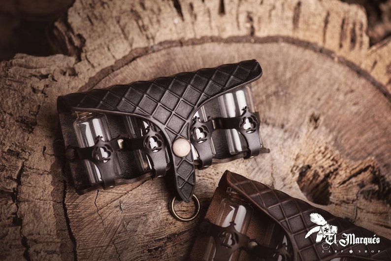 Peut inclure: &Eacute;tui en cuir noir avec un motif en losanges, contenant plusieurs petits flacons en verre. L'&eacute;tui comporte des &eacute;l&eacute;ments m&eacute;talliques d&eacute;coratifs et une fermeture avec un anneau. Le fond est une surface en bois.