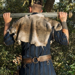 May include: A person wearing a blue tunic with a fur collar and a leather belt with metal accents. They are holding a sword over their shoulder.