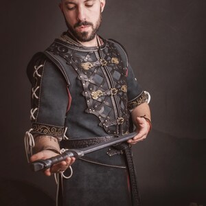 Peut inclure: Une tunique en cuir vert fonc&eacute; et marron avec des d&eacute;tails m&eacute;talliques d&eacute;coratifs et une ceinture assortie. La tunique pr&eacute;sente des coutures complexes et des fermoirs dor&eacute;s. La personne tient une dague argent&eacute;e. Le logo "El Marqu&eacute;s Larp Shop" est visible.