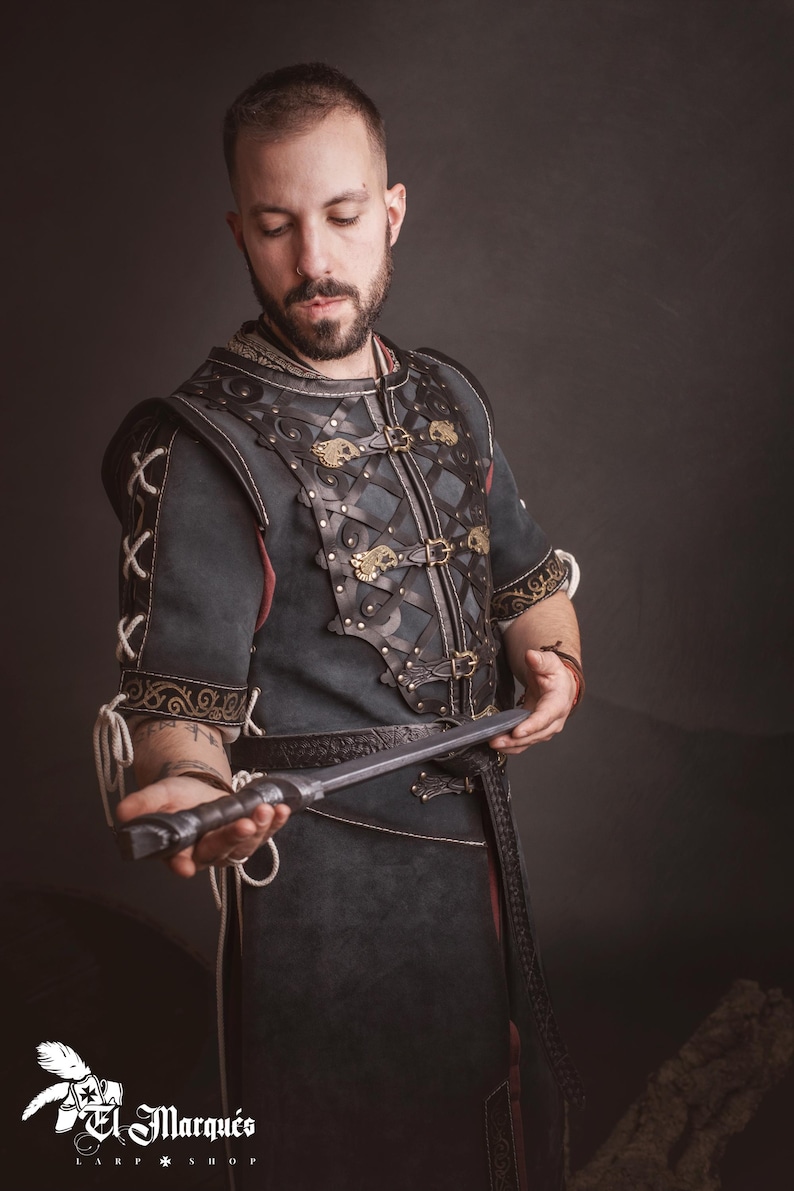 Peut inclure: Une tunique en cuir vert fonc&eacute; et marron avec des d&eacute;tails m&eacute;talliques d&eacute;coratifs et une ceinture assortie. La tunique pr&eacute;sente des coutures complexes et des fermoirs dor&eacute;s. La personne tient une dague argent&eacute;e. Le logo "El Marqu&eacute;s Larp Shop" est visible.