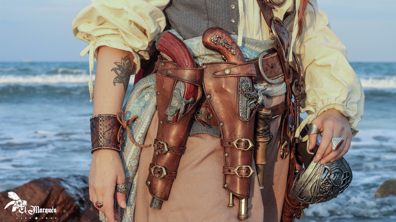 Puede incluir: Una mujer con una camisa blanca y pantalones marrones con un cintur&oacute;n de cuero marr&oacute;n. Lleva dos fundas de cuero marr&oacute;n con detalles plateados, cada una con una pistola. Tambi&eacute;n lleva una pulsera de cuero marr&oacute;n en su mu&ntilde;eca izquierda.