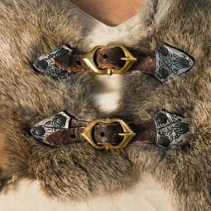 May include: Two brown leather straps with silver buckles and decorative metal accents. The straps are fastened together with a gold metal buckle.
