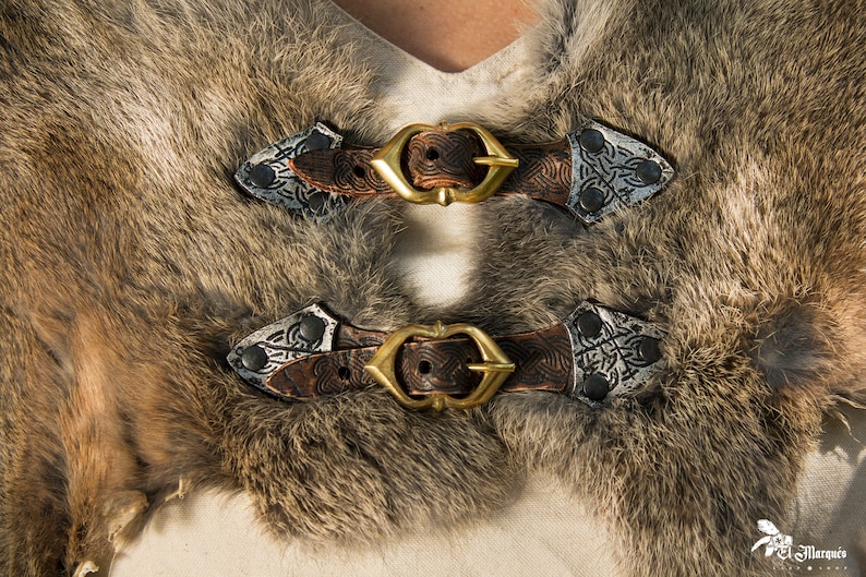 May include: Two brown leather straps with silver buckles and decorative metal accents. The straps are fastened together with a gold metal buckle.