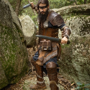 May include: A warrior in a leather costume with intricate designs, holding two swords, stands in a forest setting. The warrior has a long beard and is wearing a fur-lined helmet.