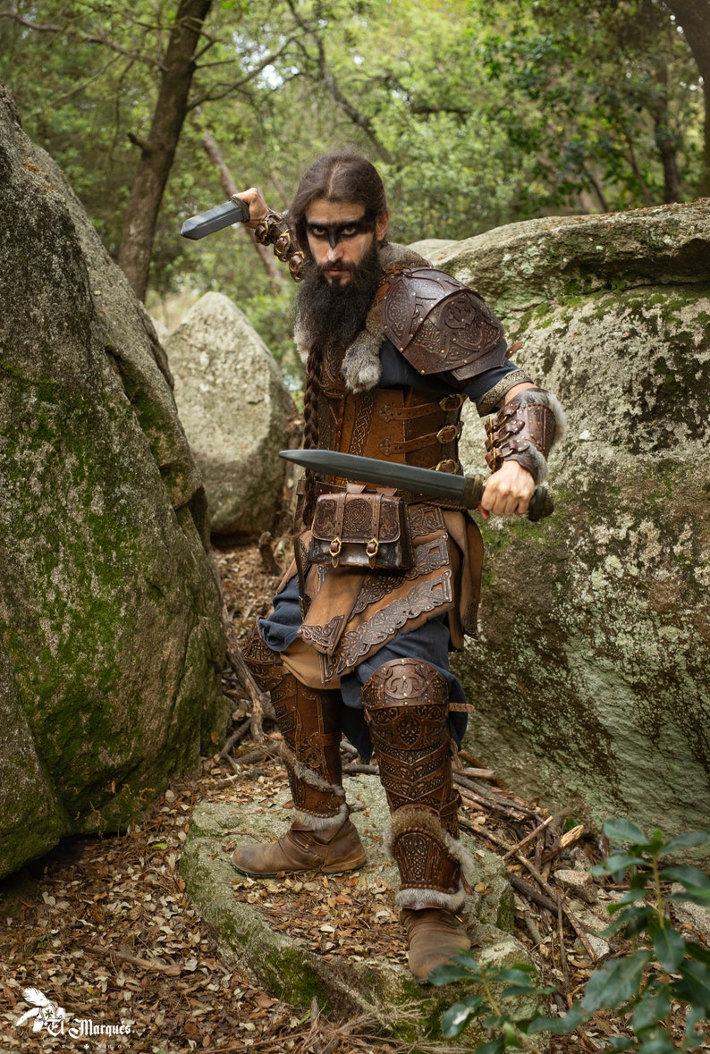 May include: A warrior in a leather costume with intricate designs, holding two swords, stands in a forest setting. The warrior has a long beard and is wearing a fur-lined helmet.