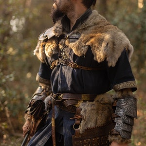 May include: A person dressed as a warrior in a forest setting. They are wearing a dark blue tunic with fur trim, leather armor, and a sword. The person has a beard and dark hair.