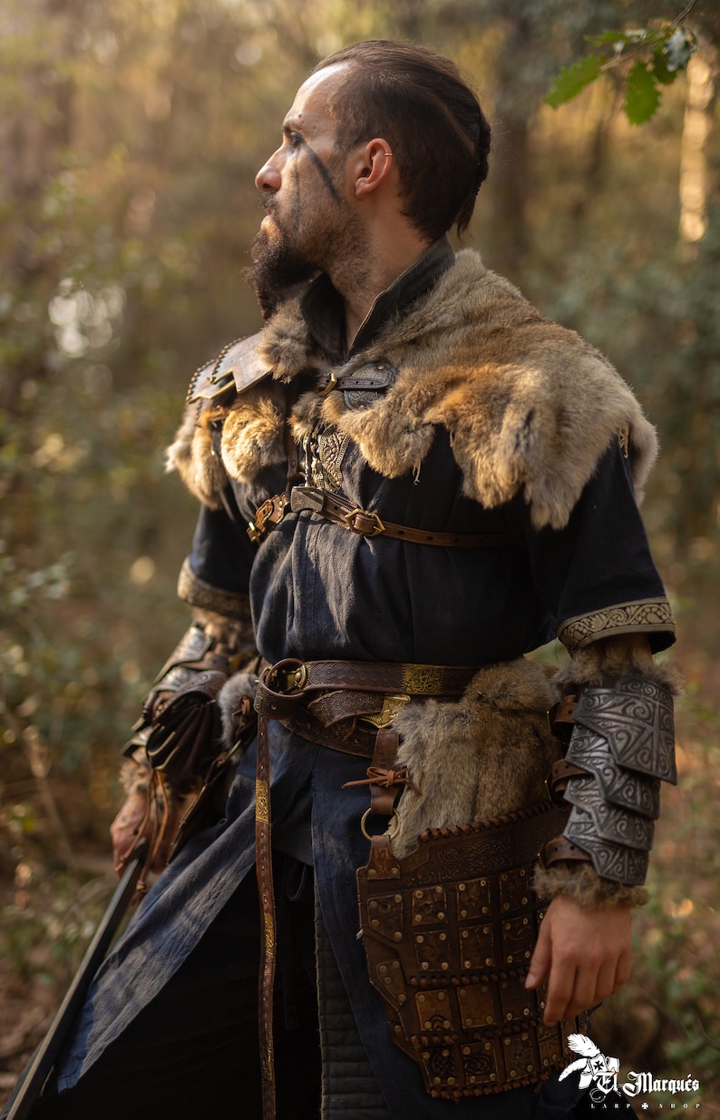 May include: A person dressed as a warrior in a forest setting. They are wearing a dark blue tunic with fur trim, leather armor, and a sword. The person has a beard and dark hair.