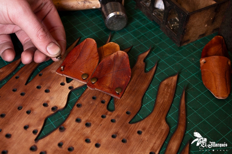 May include: A person is attaching leather pieces to a wooden board with metal rivets. The board has a flame-like design and is covered in small holes. The leather pieces are brown and have a textured surface.