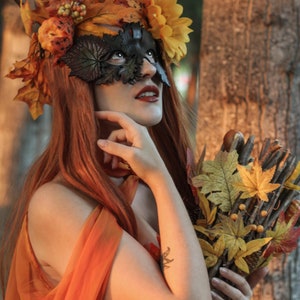 May include: A woman with long red hair wears a leafy crown and a black masquerade mask. She holds a bouquet of dried leaves and twigs in her hands.