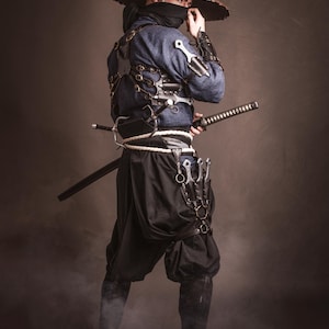 May include: A person wearing a blue and black outfit with a wide-brimmed hat, holding a sword and a variety of knives. The person is standing in a smoky environment.