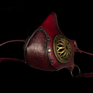 May include: A red leather mask with a gold metal design and straps. The mask has a decorative floral pattern and a circular cutout in the front.