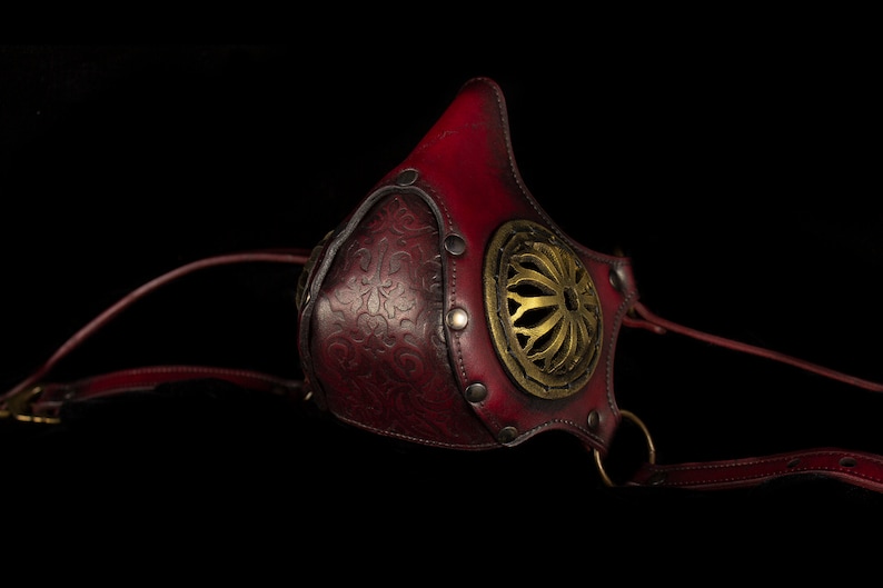 May include: A red leather mask with a gold metal design and straps. The mask has a decorative floral pattern and a circular cutout in the front.