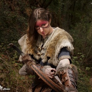 May include: A woman in a forest setting, wearing a fur-trimmed tunic and leather bracers, holds a bow and arrow. She has red and black face paint.