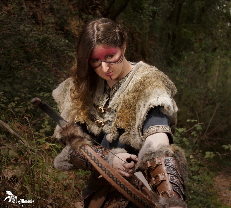 May include: A woman in a forest setting, wearing a fur-trimmed tunic and leather bracers, holds a bow and arrow. She has red and black face paint.