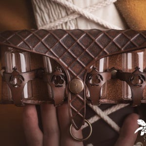 Peut inclure: Une bourse en cuir marron avec un motif en losanges, contenant trois fioles en verre. La bourse a des accents m&eacute;talliques d&eacute;coratifs et un fermoir en laiton. Le design sugg&egrave;re un th&egrave;me m&eacute;di&eacute;val ou fantastique.