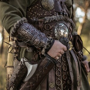 May include: A close-up of a person wearing a leather and metal armor with intricate designs. The person is holding a sword and an axe in their hands. The background is a blurred forest.