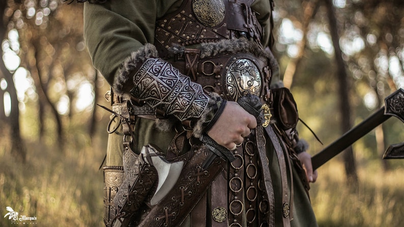 May include: A close-up of a person wearing a leather and metal armor with intricate designs. The person is holding a sword and an axe in their hands. The background is a blurred forest.