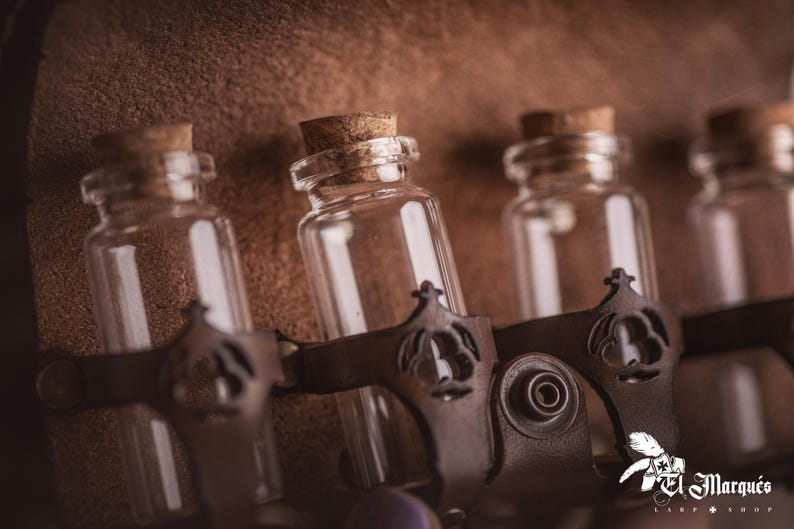 Peut inclure: Gros plan sur plusieurs petites bouteilles en verre transparent avec des bouchons en li&egrave;ge. Les bouteilles sont maintenues par des lani&egrave;res en cuir marron fonc&eacute; avec des ornements m&eacute;talliques d&eacute;coratifs. Fond textur&eacute; marron.