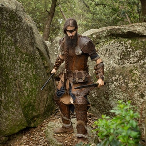 May include: A man dressed in brown leather armor with a fur-trimmed cloak and a braided beard, holding two knives in a forest setting.
