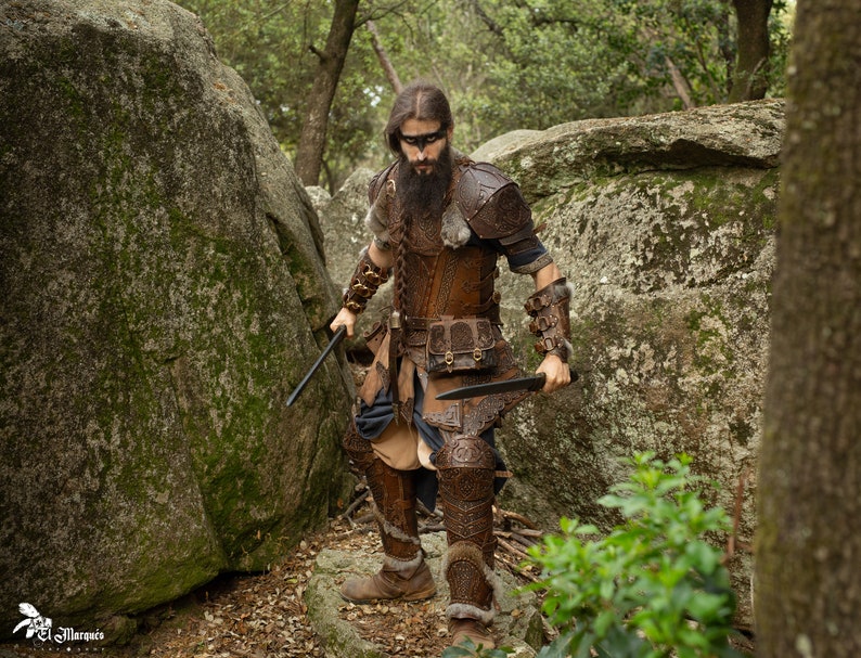 May include: A man dressed in brown leather armor with a fur-trimmed cloak and a braided beard, holding two knives in a forest setting.