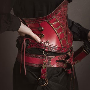 K&ouml;nnte beinhalten: Ein rotes Lederkorsett mit aufwendigen floralen Mustern und Metallverzierungen. Das Korsett wird &uuml;ber einem schwarzen Hemd getragen und mit einem passenden roten Lederg&uuml;rtel und einer Schwertscheide kombiniert.