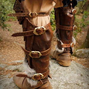 May include: Brown leather leg armor with fur trim and multiple buckles. The armor is worn on both legs and has a distressed look.