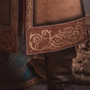 Peut inclure: Gros plan sur un v&ecirc;tement en cuir marron avec une bordure dor&eacute;e orn&eacute;e de n&oelig;uds celtiques. Le v&ecirc;tement semble faire partie d'un costume, avec des coutures visibles et une botte en cuir marron fonc&eacute;. L'image a une tonalit&eacute; chaude et terreuse.