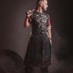 Peut inclure: Une personne portant une tunique noire et rouge de style m&eacute;di&eacute;val avec des d&eacute;tails dor&eacute;s et une jupe assortie. La tunique pr&eacute;sente des d&eacute;tails complexes et est associ&eacute;e &agrave; des bottes noires. La personne tient une massue en bois sur son &eacute;paule.