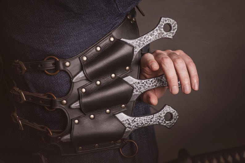 May include: A leather belt with four metal buckles and four sheaths for holding throwing knives. The knives are silver with a textured finish.