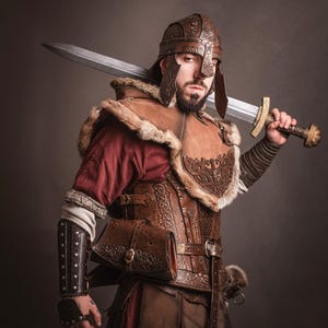 K&ouml;nnte beinhalten: Ein Krieger in brauner Lederr&uuml;stung, einem passenden Helm und einem pelzgef&uuml;tterten Umhang, der ein gro&szlig;es Schwert h&auml;lt. Die R&uuml;stung weist aufwendige gepr&auml;gte Designs auf. Der Krieger tr&auml;gt ein rotes Hemd unter der R&uuml;stung.