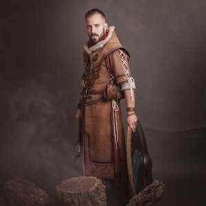 Peut inclure: Une personne porte un costume en cuir marron avec une capuche bord&eacute;e de fourrure, tenant un bouclier. Le costume pr&eacute;sente des coutures et des d&eacute;tails complexes. Le bouclier est bleu et noir. L'image est sur un fond sombre et enfum&eacute;.