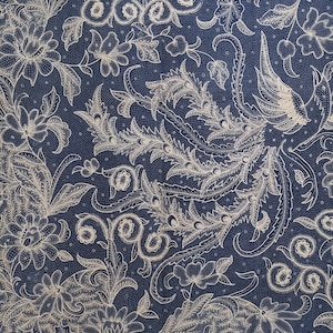 Op de afbeelding: Een marineblauwe stof met een gedetailleerd bloemenpatroon in gebroken wit. Het ontwerp toont gestileerde bloemen, bladeren en wervelende ranken, wat een rijke, getextureerde uitstraling creëert. De achtergrond heeft een subtiele gestippelde textuur.