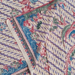 Può includere: Primo piano di un tessuto fantasia con sfondo crema. Il tessuto presenta un motivo ripetuto di fiori stilizzati, foglie e motivi a onde in tonalità blu, viola e rosso. I bordi sono rifiniti con bordi decorativi.