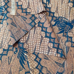 Op de afbeelding: Een close-up van een blauwe en bruine stof met een herhalend patroon van gestileerde bladeren en bloemen. De stof is op verschillende plaatsen gevouwen, waardoor het patroon eronder zichtbaar wordt.