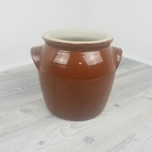 May include: A brown ceramic pot with a wide opening and two small handles. The interior is a light cream color. The pot has a glossy finish and is sitting on a light gray wooden surface. This decorative pot is suitable for kitchen or home decor.