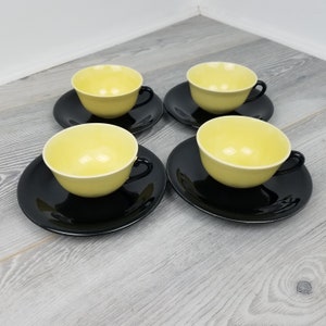 May include: A set of four tea cups and saucers. The cups are pale yellow with black handles, sitting on black saucers. The set is displayed on a light grey wooden surface.
