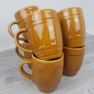 May include: A set of six brown ceramic mugs shaped like barrels. The mugs have a wood grain pattern and are stacked on top of each other.