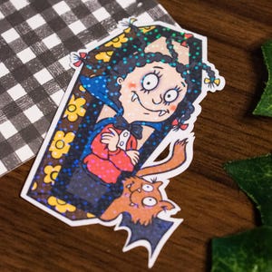 Könnte beinhalten: Ein farbenfroher Aufkleber mit einer Cartoon-Vampirfigur in einem Sarg, mit einer Fledermaus. Der Vampir hat schwarzes Haar, ein rotes Outfit und einen blauen Umhang. Gelbe Blumenakzente und ein schwarz-weißes Karomuster befinden sich im Hintergrund.
