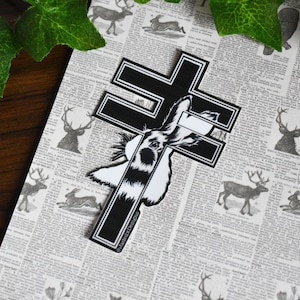 May include: Black and white sticker of a rabbit with a cross behind it. The sticker is on a newspaper background with images of deer and rabbits.