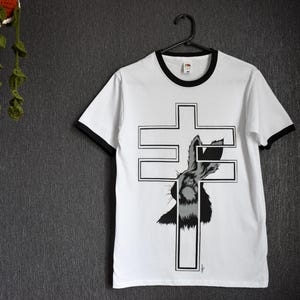 Puede incluir: Camiseta blanca con ribetes negros y un diseño gráfico de un conejo dentro de una cruz. La camiseta está colgada en una percha negra. Una planta de ganchillo cuelga en la esquina superior izquierda.