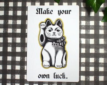 May include: A white card featuring a black and white illustration of a Maneki-neko cat, with the text "Make your own luck." The cat is depicted in a seated pose, with one paw raised. The background is a monochrome checkered pattern.