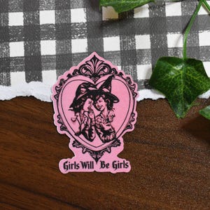 May include: A pink sticker featuring a black illustration of two women in witch hats within a heart-shaped frame. The text "Girls Will Be Girls" is at the bottom. The sticker is on a wooden surface with a checkered pattern in the background.